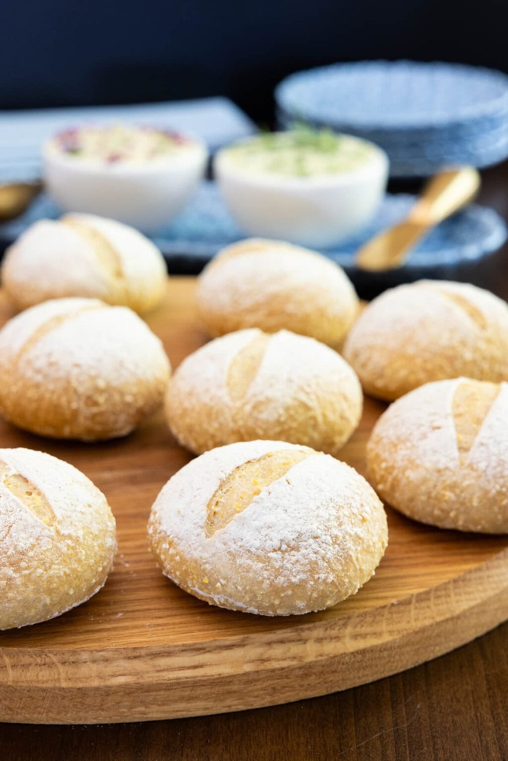 Close-up of homemade no-knead Italian-style dinner rolls with flour-dusted tops on a wooden serving board, with compound butters softly blurred in the background.