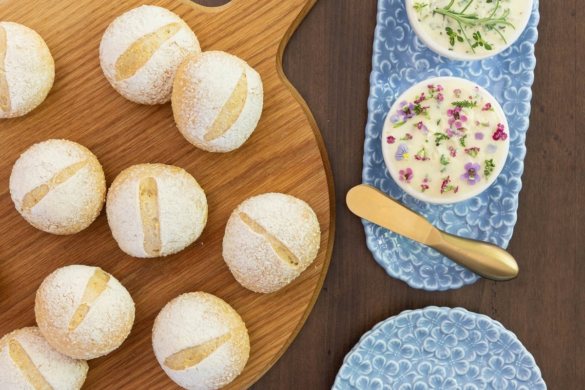 Overhead view of homemade no-knead Italian-style dinner rolls on a wooden serving board with herb butter and flower-topped compound butter on a blue serving tray.
