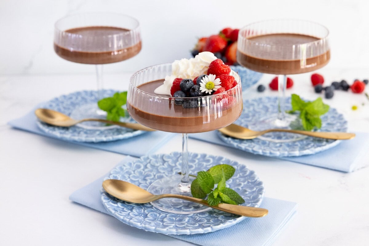 Chocolate panna cotta served in elegant coupe glasses with whipped cream, fresh raspberries, blueberries and mint on blue patterned dessert plates.