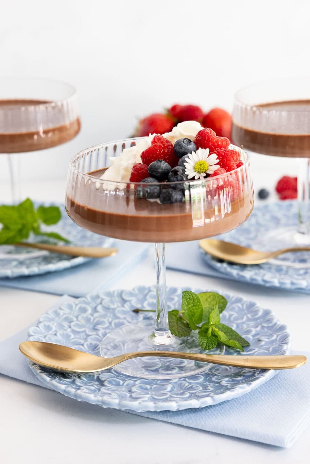 Chocolate panna cotta in an elegant coupe glass topped with whipped cream, fresh raspberries and blueberries, served on a blue patterned dessert plate.