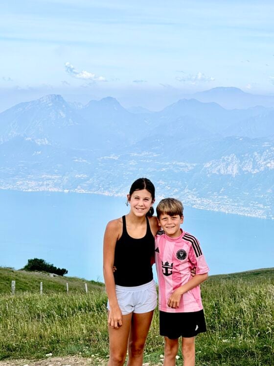 Two grandchildren smiling on Monte Baldo with panoramic views of Lake Garda and the surrounding mountains in Italy.