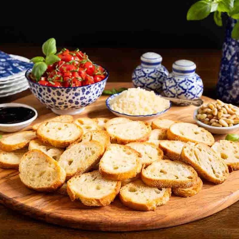 Tomato bruschetta in a blue and white bowl on a large rounds board with crostini, and small bowls of balsamic and parmesan.