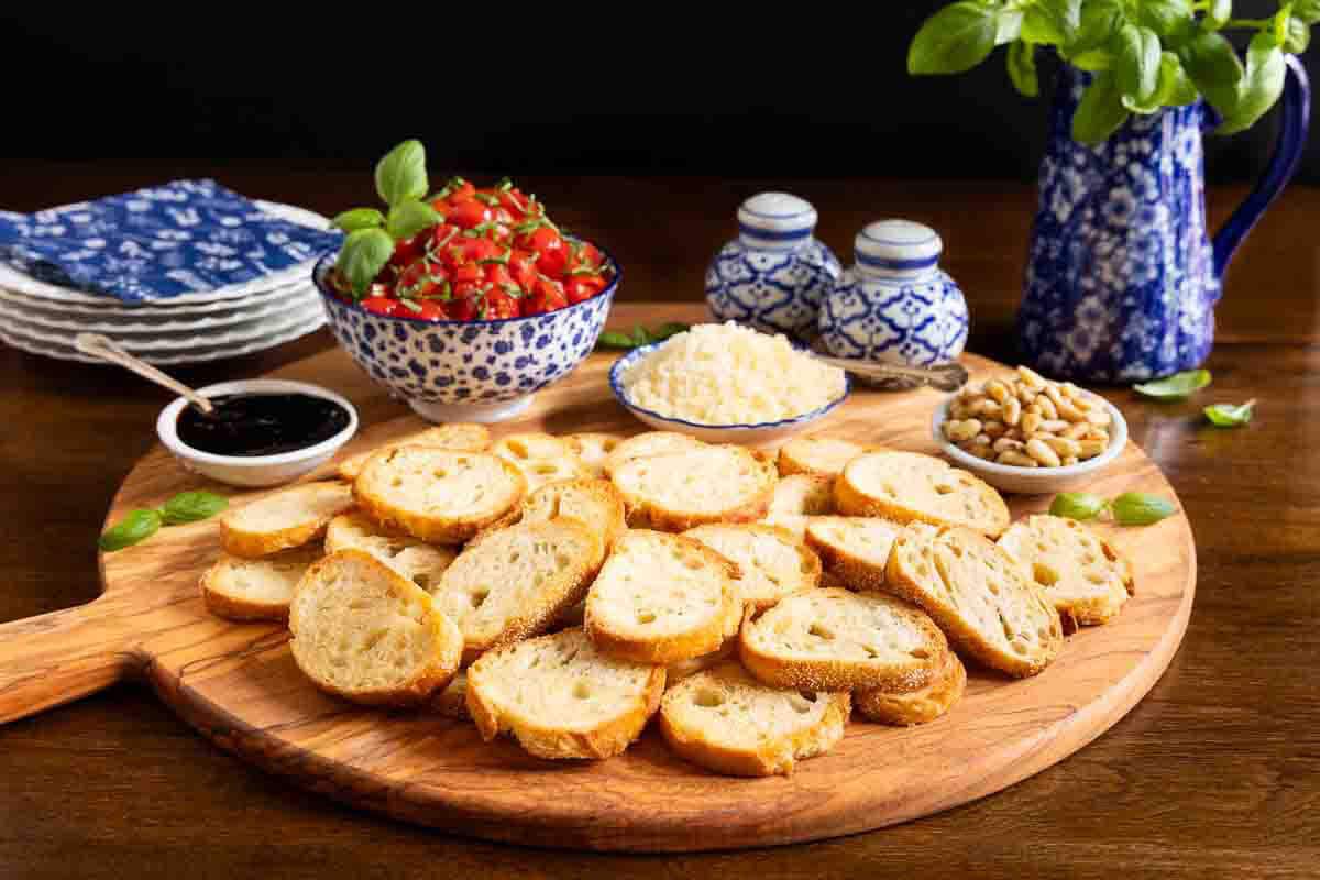 Tomato bruschetta in a blue and white bowl on a large rounds board with crostini, and small bowls of balsamic and parmesan.