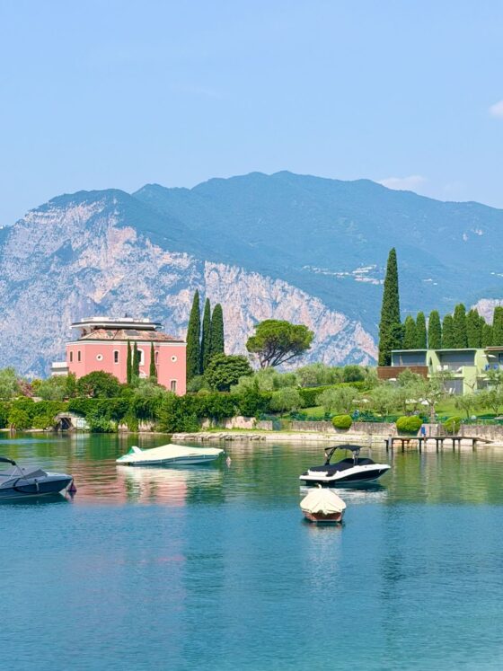 Boats floating on the calm waters of Lake Garda with colorful villas, cypress trees, and dramatic mountains in the background.