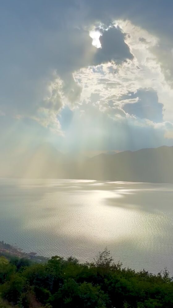 Sunbeams breaking through dramatic storm clouds over Lake Garda, illuminating the shimmering water below.