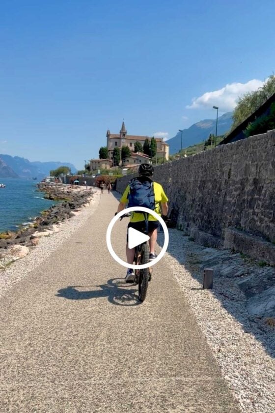 Scott riding a bicycle along a scenic lakeside bike path beside Lake Garda with mountains, blue water, and a historic church in the background.