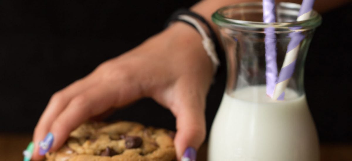 A shot of a young girl's hands sneaking a One Bowl Toffee Bar Chocolate Chip Cookie from a stack next to a carafe of milk.