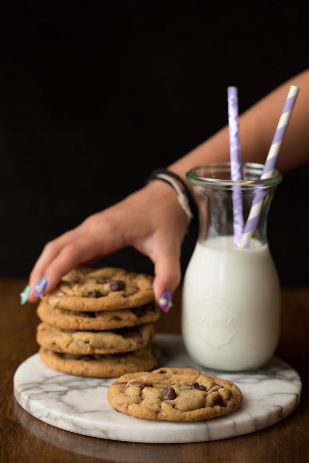 A shot of a young girl's hands sneaking a One Bowl Toffee Bar Chocolate Chip Cookie from a stack next to a carafe of milk.