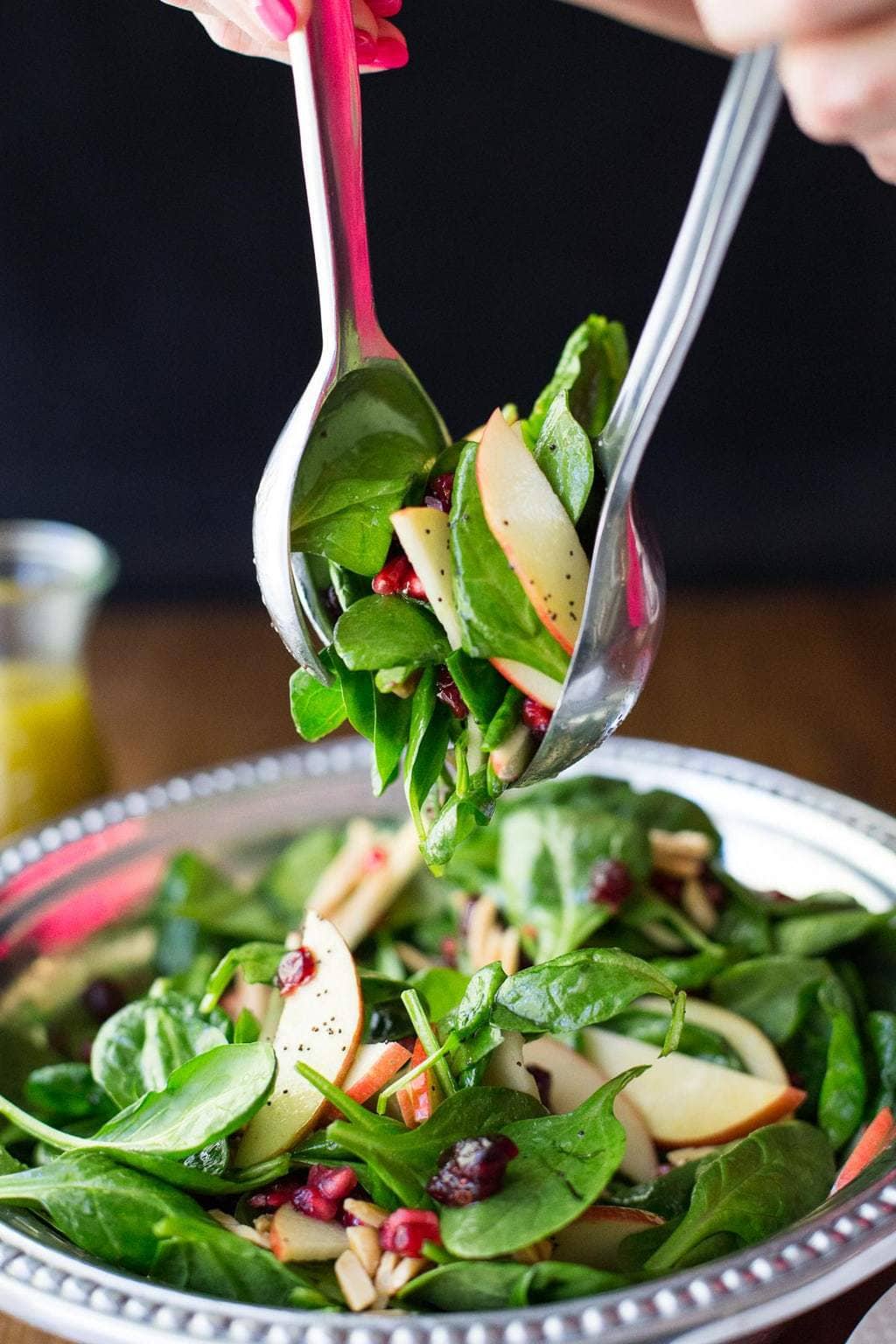 Vertical closeup photo of a Apple Cranberry Spinach Salad with salad serving utensils being used to toss the salad.