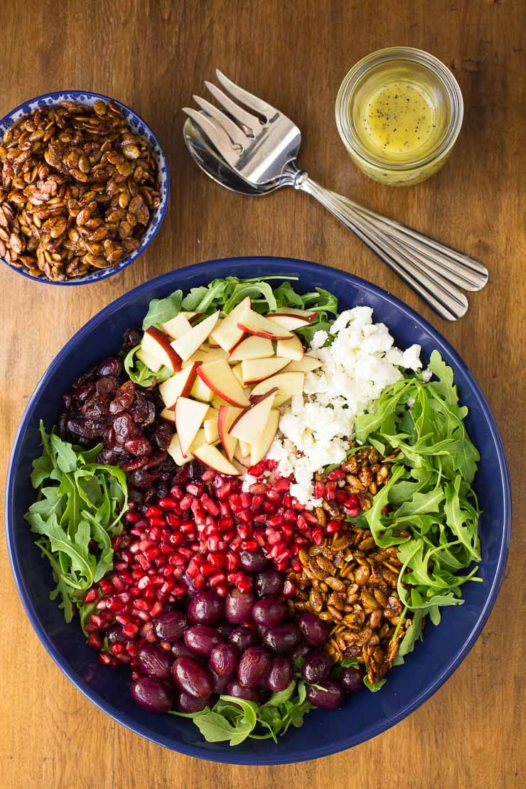 Overhead photo of a serving bowl of Arugula Roasted Grape Salad prior to being tossed together. Surrounding the bowl are serving utensils, a carafe of dressing and a smaller dish of candied pumpkin seeds.
