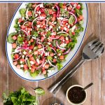 Overhead vertical photo of an Asian Watermelon Herb Salad on a wood table surrounded by fresh herbs and a bowl of slivered almonds.