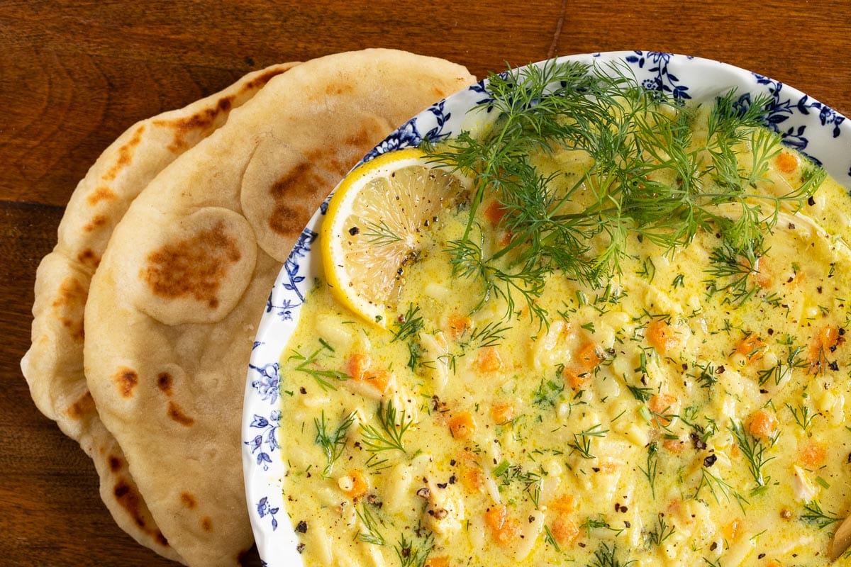 Horizontal overhead photo of a serving bowl of Avgolemono (Greek Chicken Pasta Soup) with a piece of homemade pita bread along side on a wood table.
