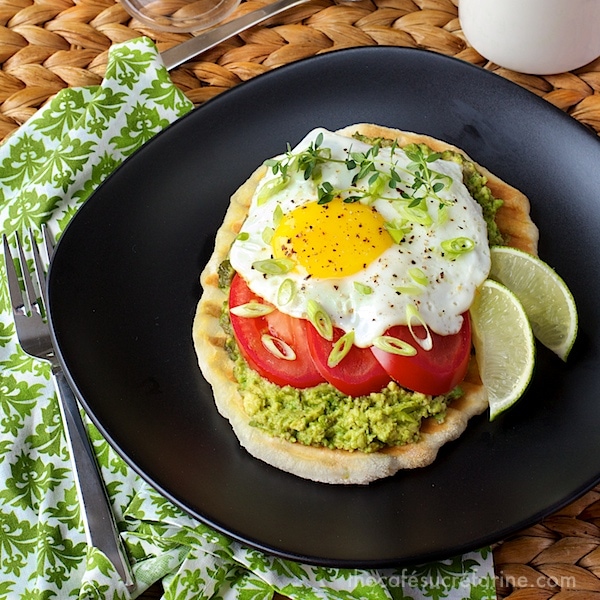 Photo of a black plate with Avocado Breakfast Flatbreads on it on a wicker table and a green and white patterned napkin underneath the plate.