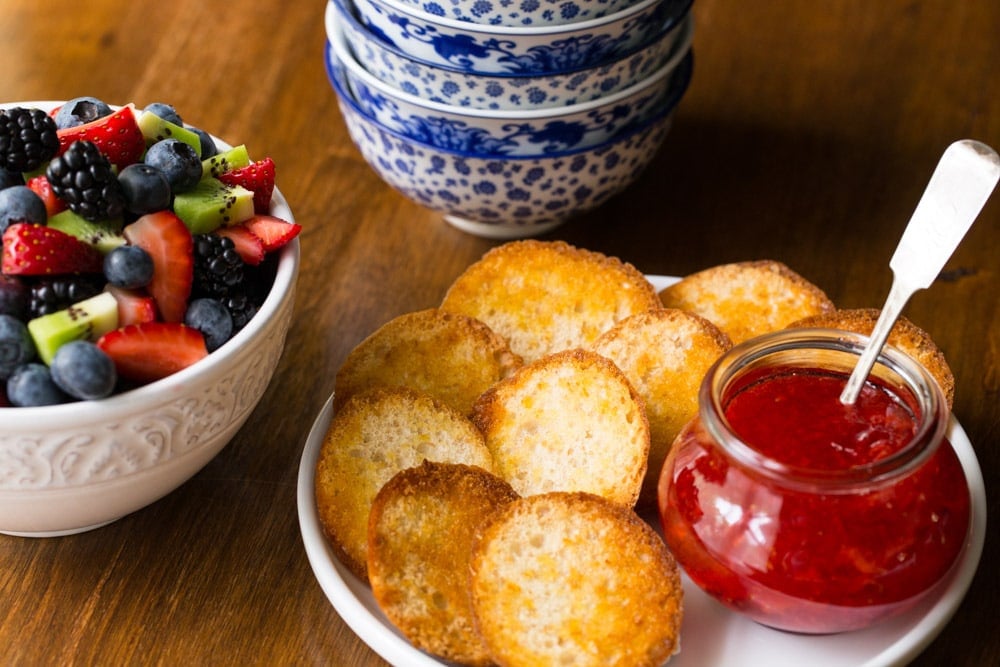 Photo of the tops of Baked Eggs in Bread Baskets served with jam and a bowl of fresh fruit.