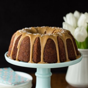 Vertical picture of Banana Pound Cake with Salted Toffee Icing on a light blue cake stand with white plates stacked in the background