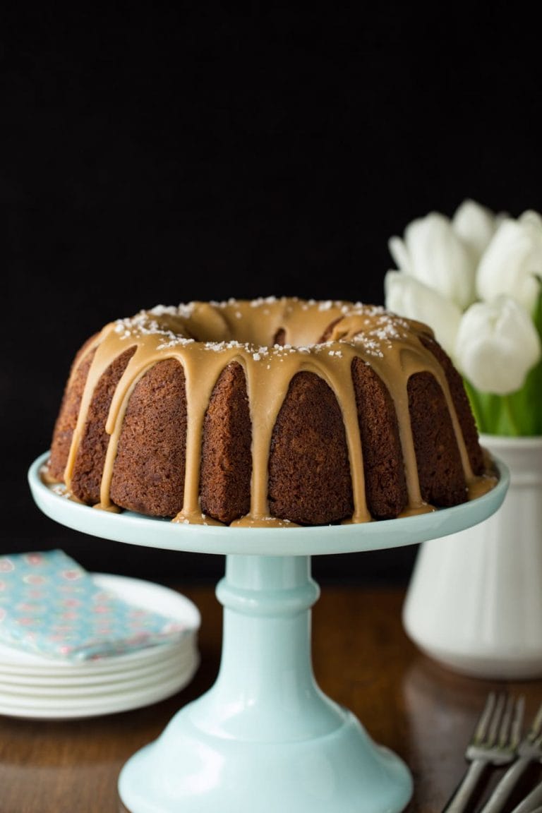 Vertical picture of Banana Pound Cake with Salted Toffee Icing on a light blue cake stand with white plates stacked in the background