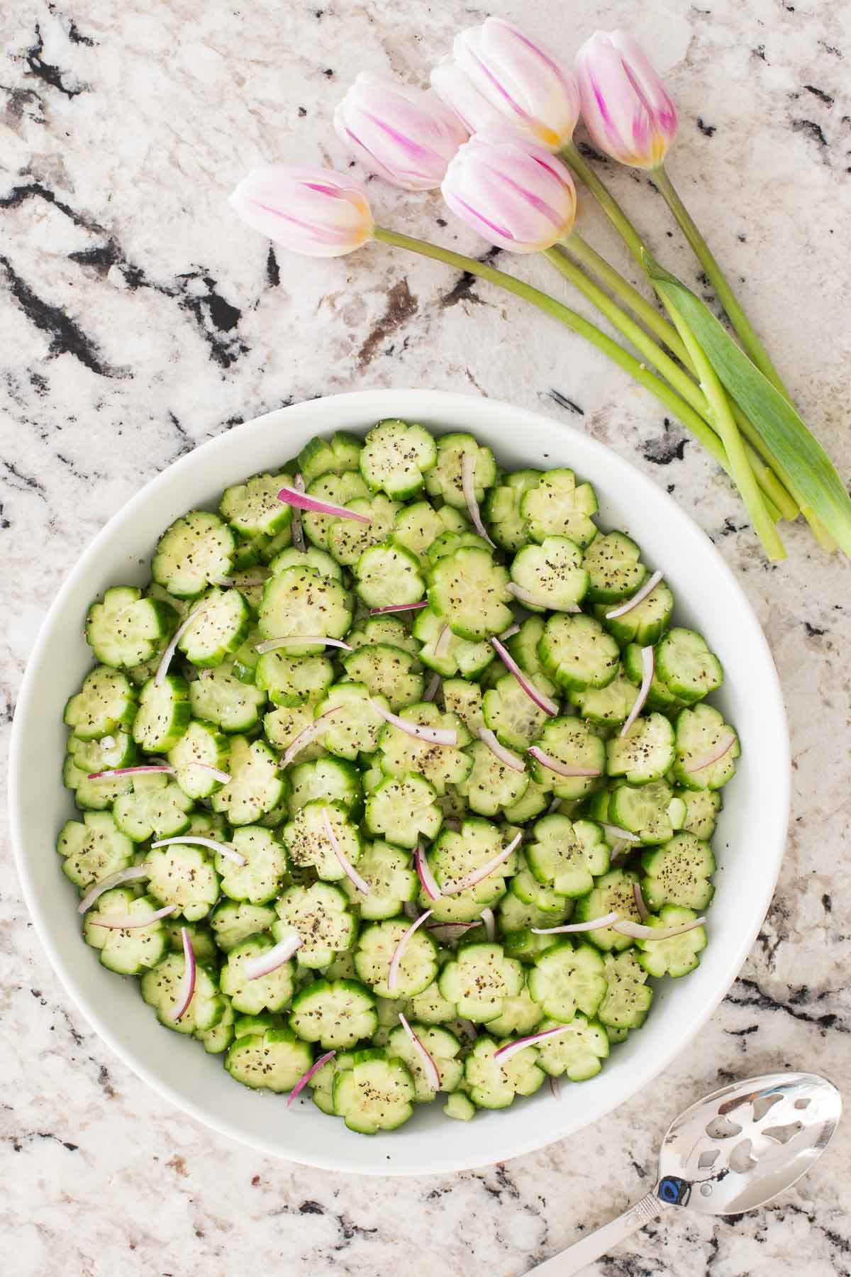 Vertical overhead photo of a white bowl of Cucumber Flowers on a marble surface.