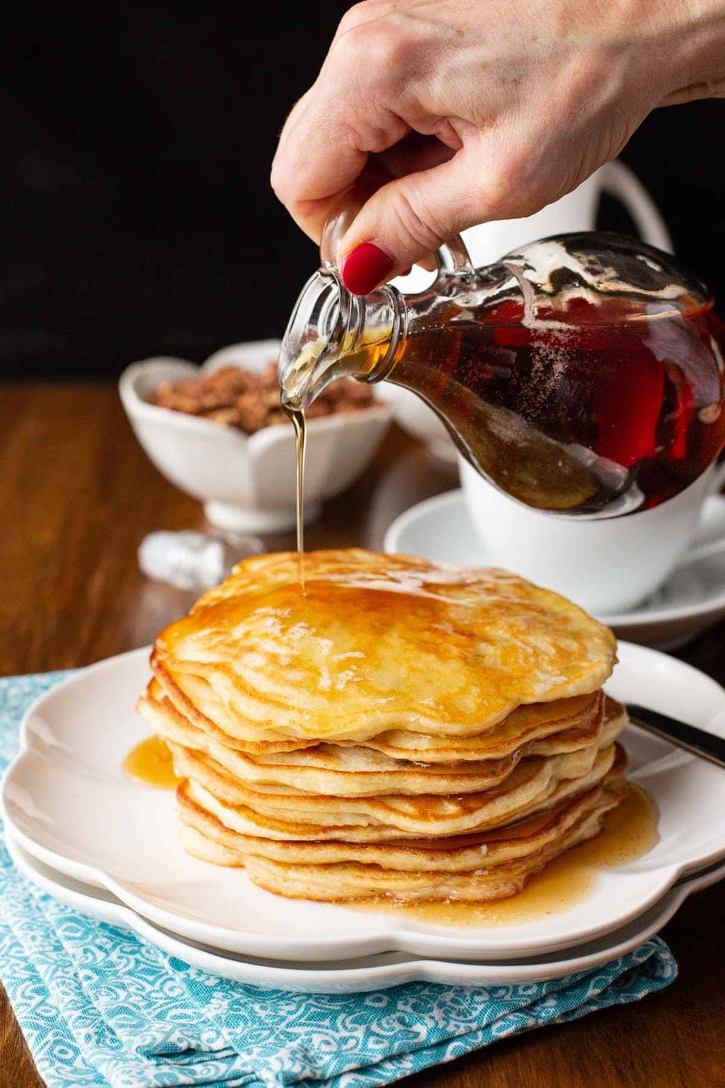 Vertical photo of a stack of Best Ever Pancakes (with Greek yogurt) on a white serving plate with a hand pouring maple syrup over the pancakes.