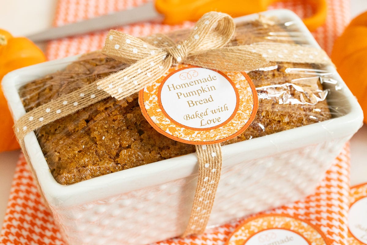 Vertical closeup photo of a loaf of Better Than Starbucks Pumpkin Bread (One-Bowl, No-Mixer!) in a decorative white baking pan wrapped up for gift giving with a custom label on top.