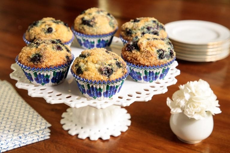 Horizontal closeup photo of a batch of Better than Starbucks Blueberry Muffins on a white lattice pedestal serving plate.
