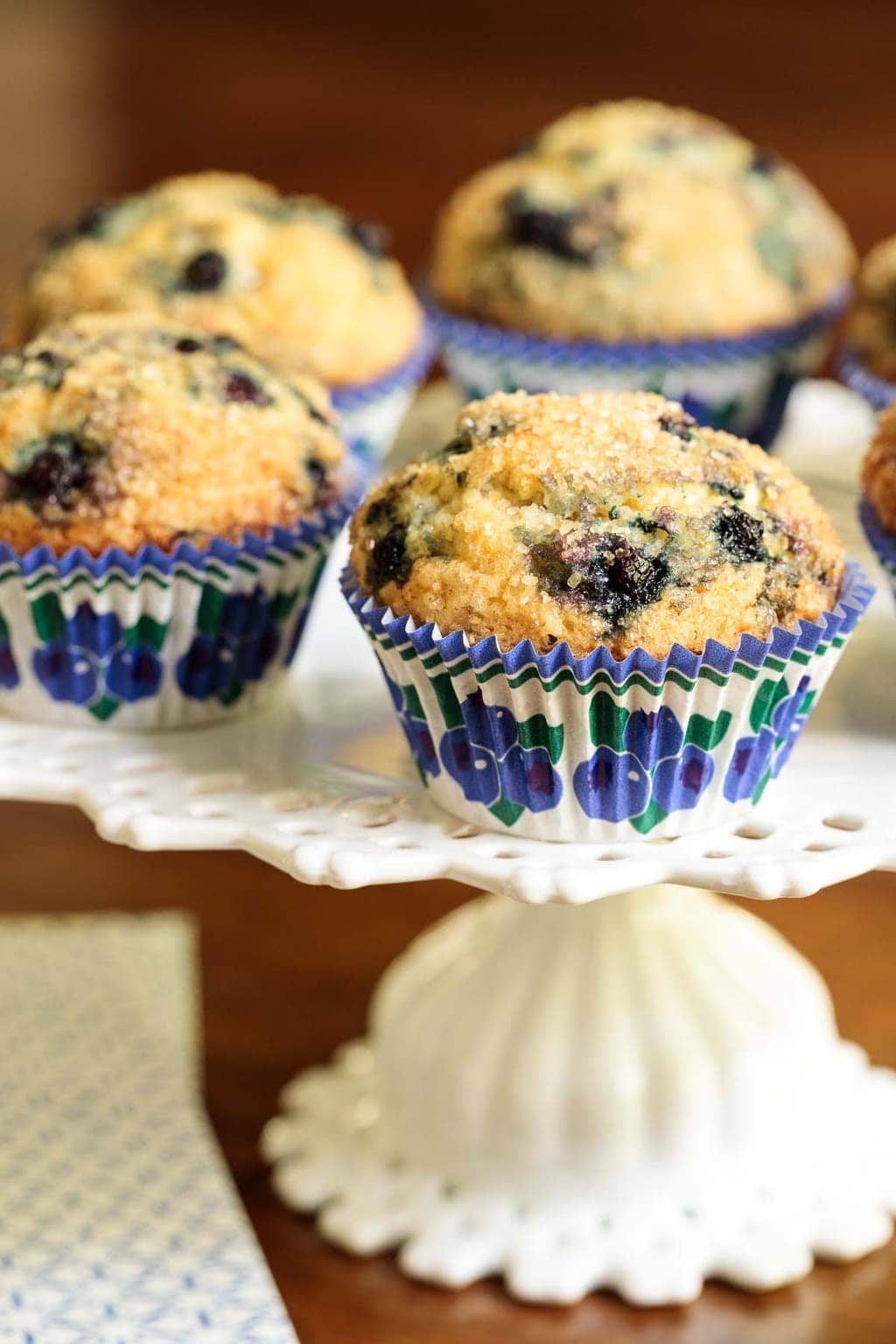 Vertical closeup photo of Copycat Starbucks Blueberry Muffins on a white cake stand resting on a wood table.