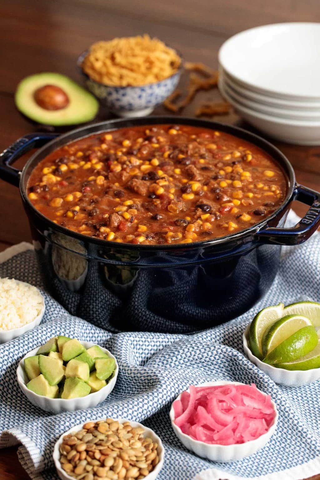 Photo of a large Staub pot of Instant Pot Black Bean Beef Tortilla Soup surrounded by small white bowls of garnish ingredients and an avocado sliced in half.
