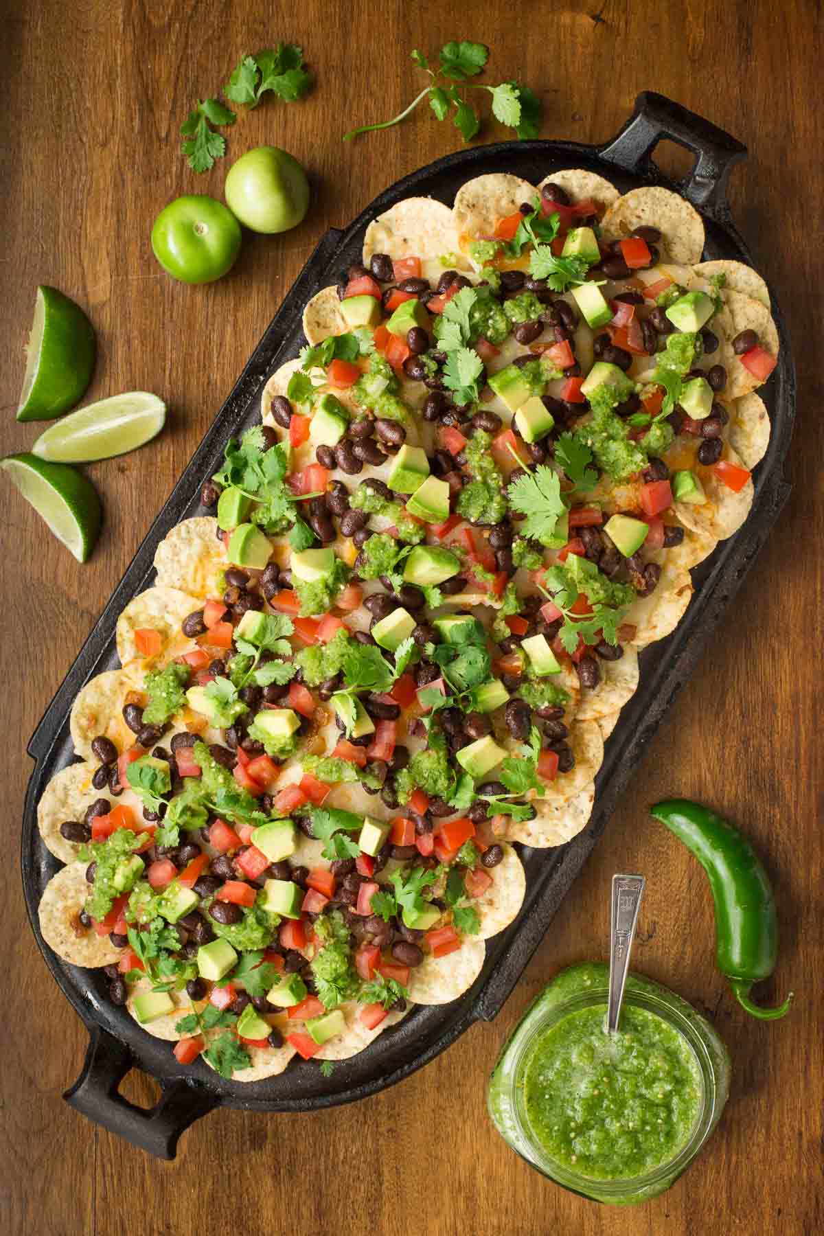 Overhead vertical photo of Black Bean Nachos with Salsa Verde in a cast iron baking dish on a wooden table.