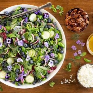 Horizontal overhead photo of a white serving bowl of Blueberry Arugula Herb Salad on a wood table.