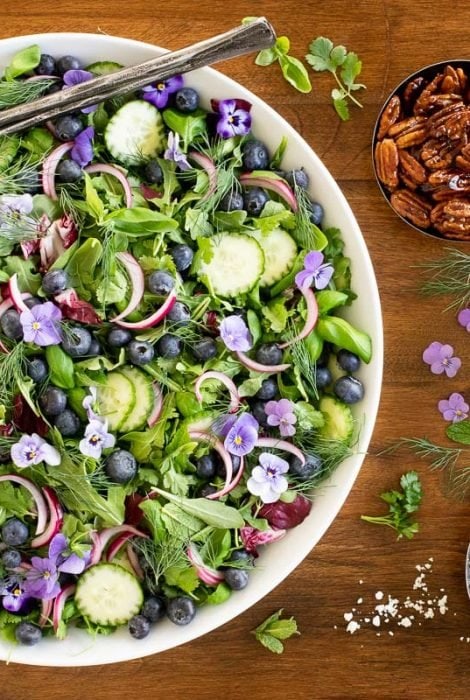Horizontal overhead photo of a white serving bowl of Blueberry Arugula Herb Salad on a wood table.