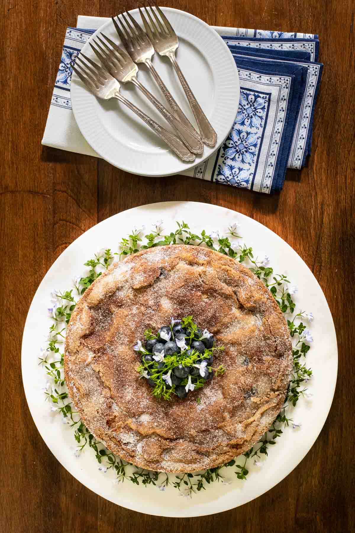 Overhead vertical photo of a Blueberry Buttermilk Breakfast Cake on a white serving plate with forks and napkins.