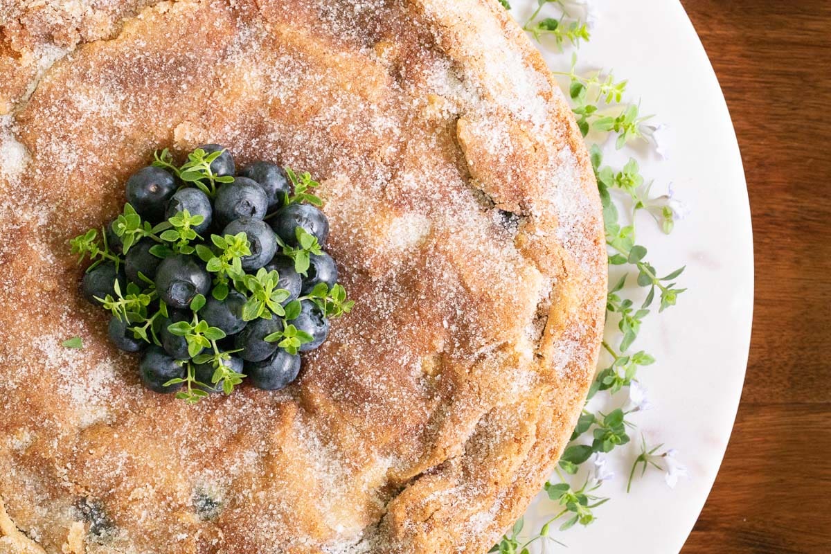 Overhead vertical photo of the top of a Blueberry Buttermilk Breakfast Cake garnished with fresh blueberries and herbs.
