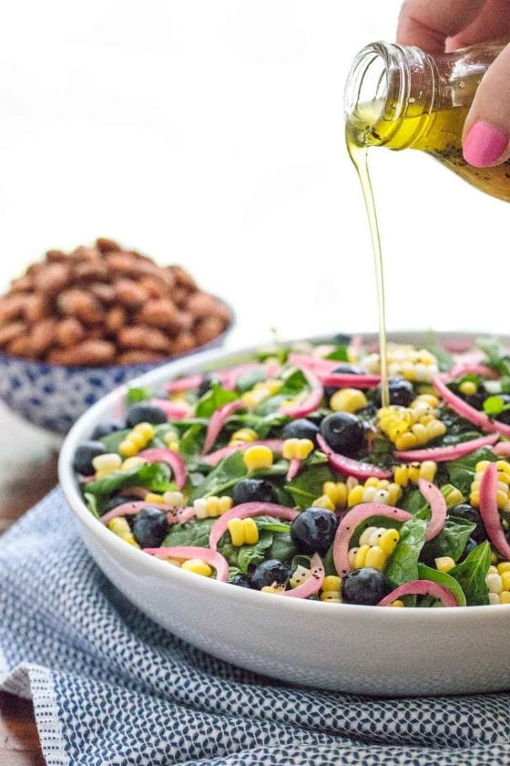Vertical photo of Blueberry and Fresh Corn Spinach Salad in a white serving bow with dressing being poured on top of the salad.
