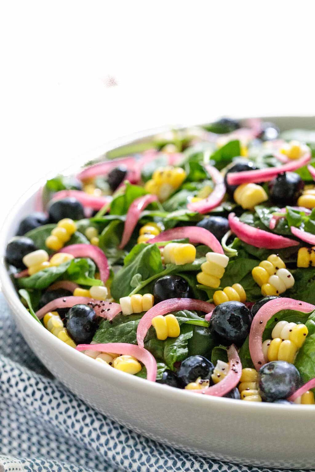 Vertical photo of a Blueberry and Fresh Corn Spinach Salad in a white serving dish with a blue and white patterned cloth in the foreground.