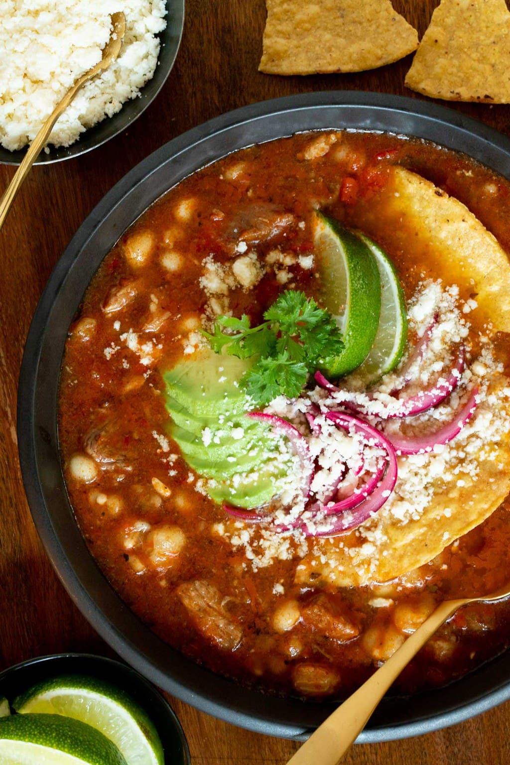 Vertical overhead photo of a bowl of Braised Pork Shoulder Pozole garnished with lime wedges, corn tortillas, sliced avocado, red onions and cilantro.