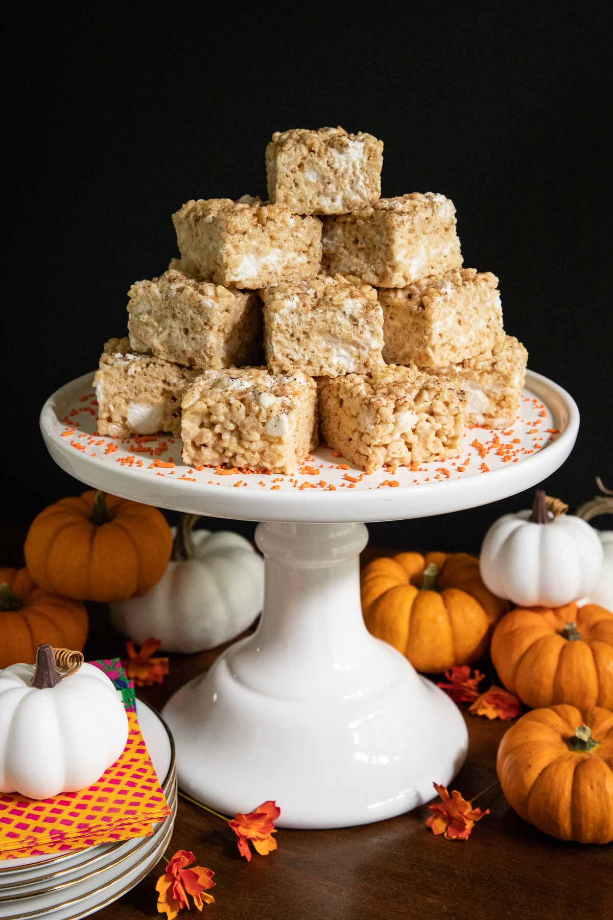 Vertical photo of a stack of Pumpkin Spiced Brown Butter Rice Krispie Treats on a white pedestal stand surrounded by fall mini pumpkins.