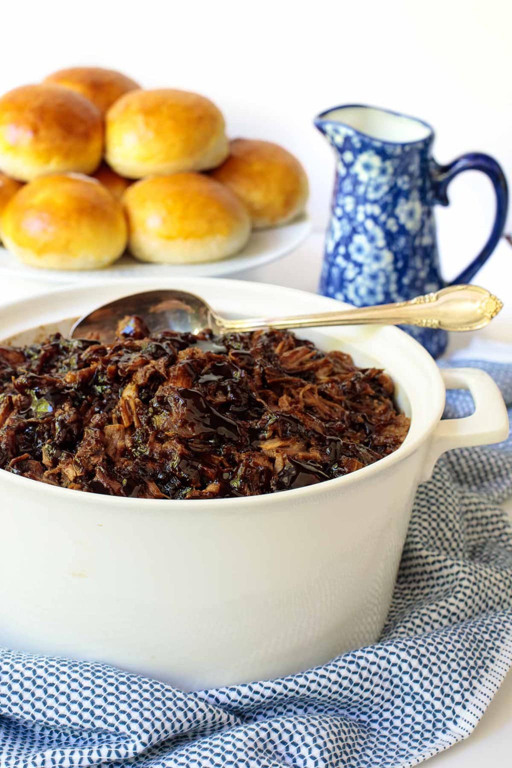 Photo of a white serving dish filled with Brown Sugar Balsamic Pulled Pork. The dish is on a blue and white patterned serving cloth and homemade brioche buns are on a plate in the background.