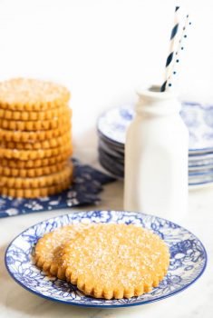 Vertical photo of two Brown Sugar Honey Shortbread Cookies on a plate with a stack of the cookies and a pint of milk in the background.