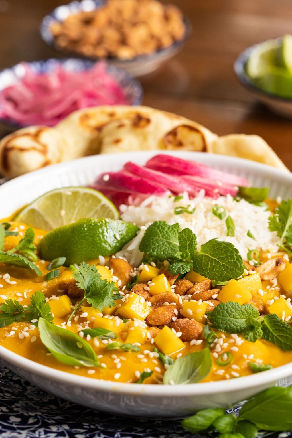 Closeup photo of a white serving bowl of Butternut Chicken Coconut Curry with naan bread, peanuts and red onions in the background.