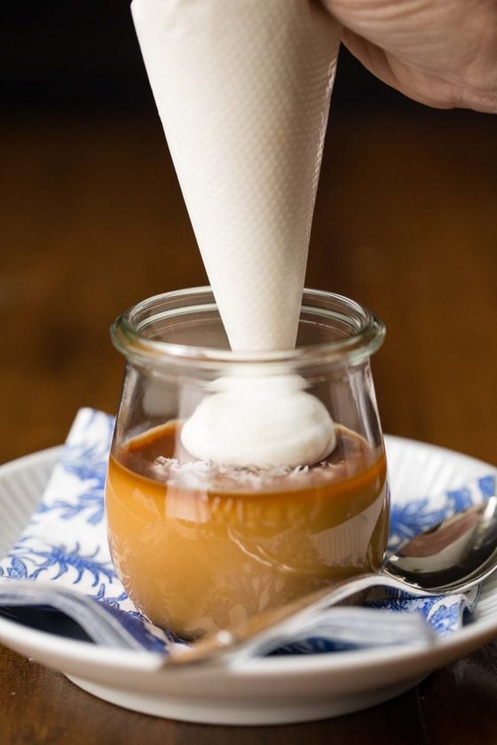 Photo of a person filling the top of a glass jar of Butterscotch Pots de Créme with whipped cream.