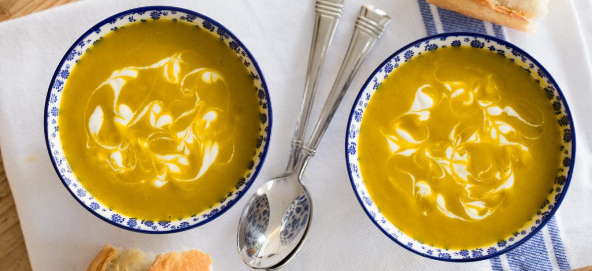 Overhead photo of two bowls of Carrot Coriander Soup on a white and blue napkin with spoons and crusty French bread nearby.