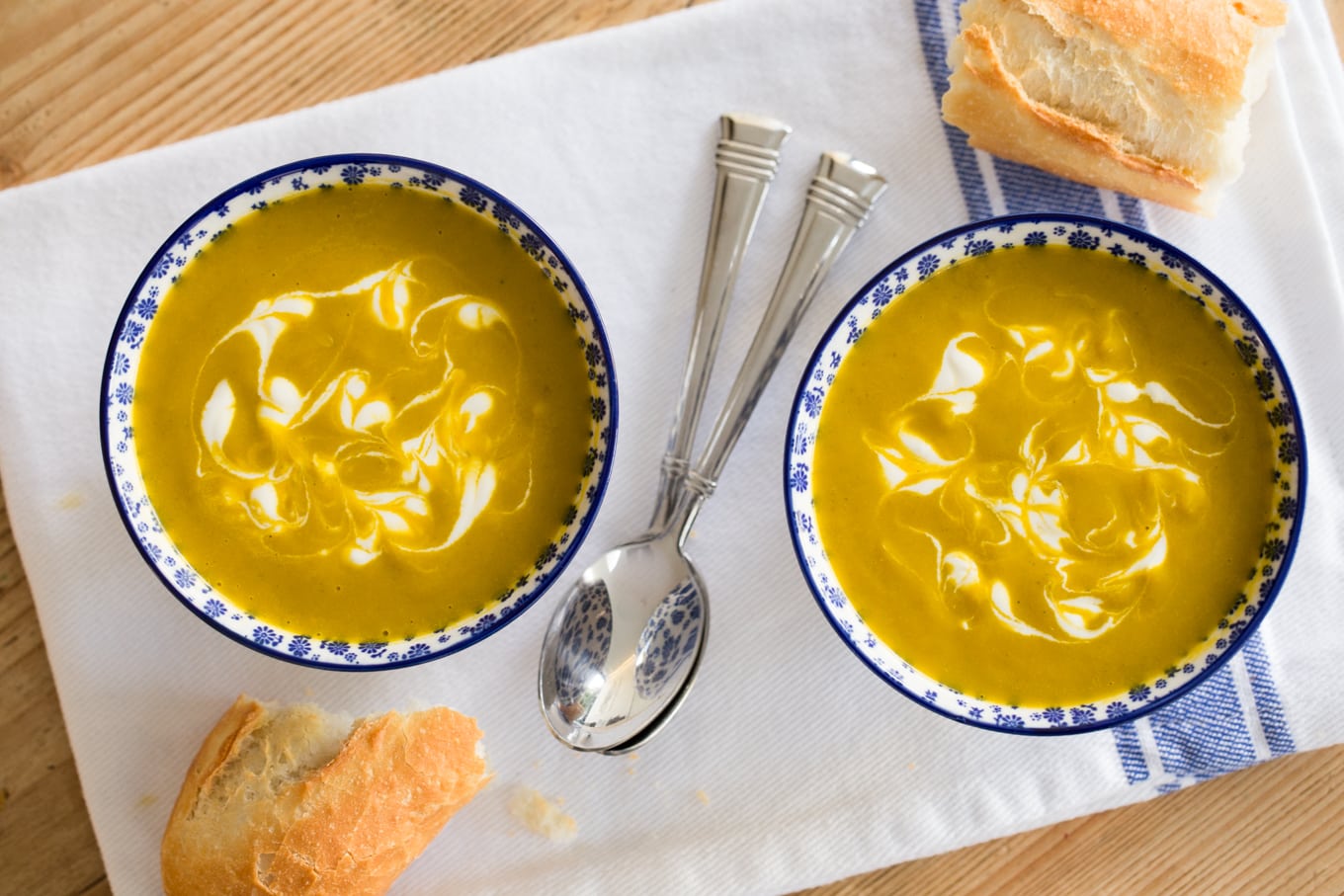 Overhead photo of two bowls of Carrot Coriander Soup on a white and blue napkin with spoons and crusty French bread nearby.