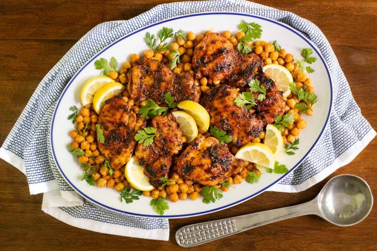 beautifully charred grilled chicken served with chickpeas, lemon wedges, and fresh herbs on a white platter.
