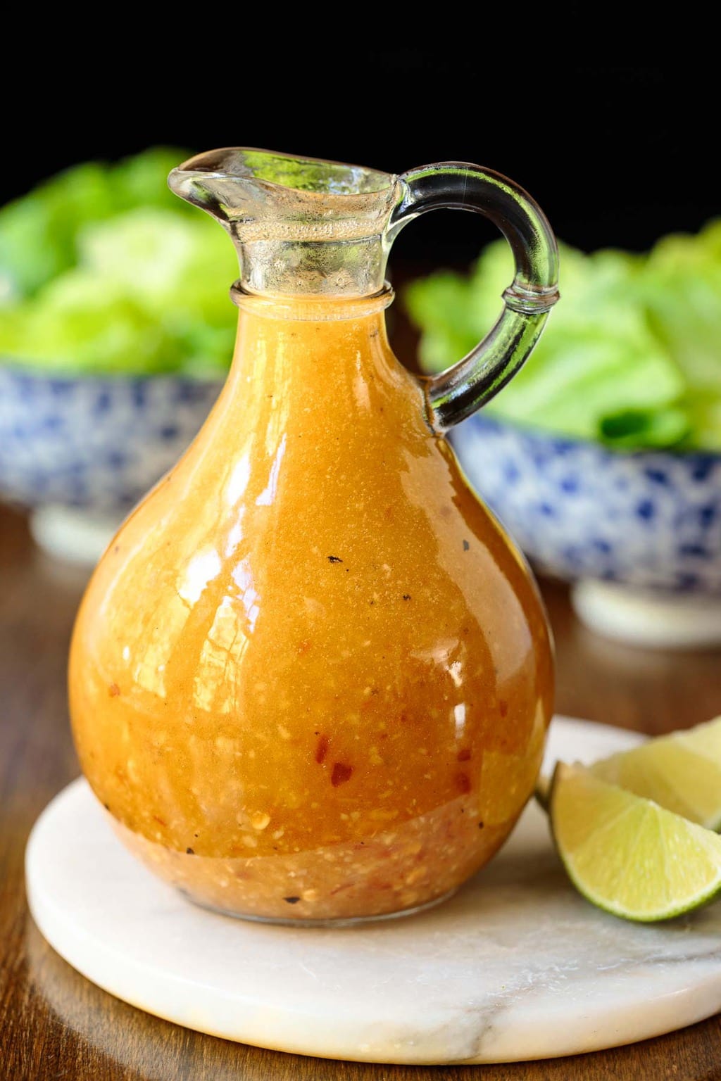 Photo of a small glass pitcher of Chili Lime Salad Dressing on a marble surface with fresh garden salads in the background.