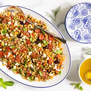 Horizontal overhead photo of a Chopped Mediterranean Farro Salad on a white oval serving platter surrounded by basil and dill garnishes.