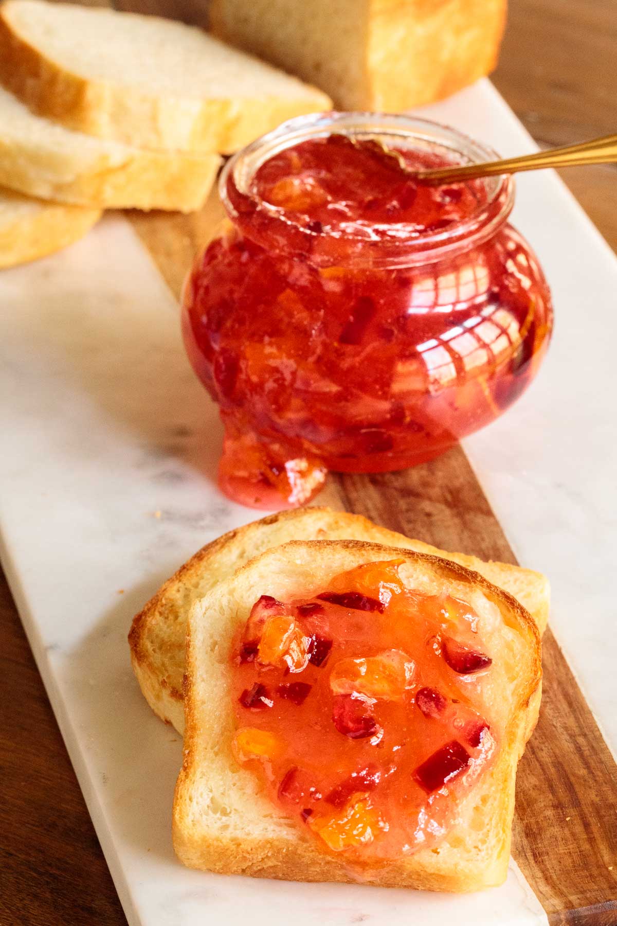 Photo of Easy Orange-Cranberry Christmas Marmalade spread on fresh brioche bread on a marble and wood serving platter with a jar of the marmalade in the background.