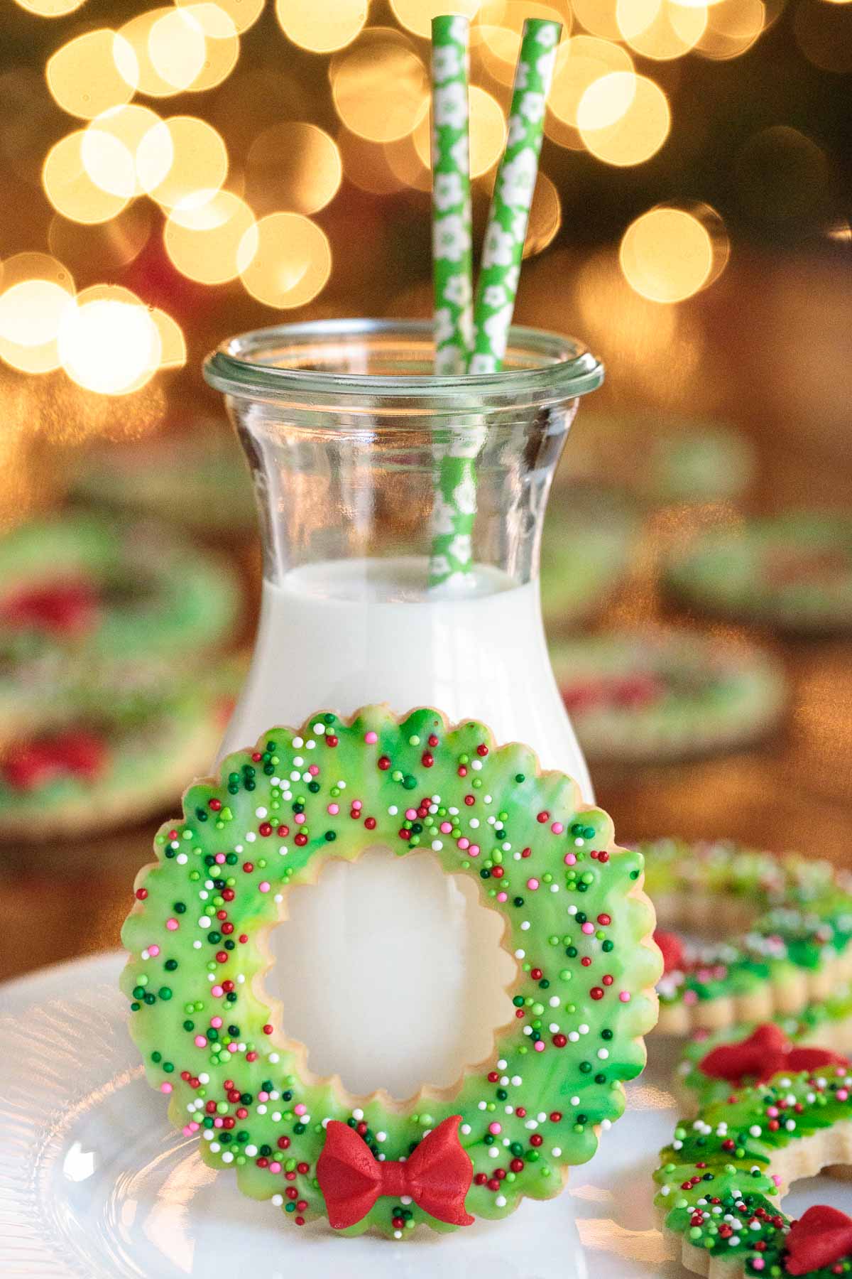 Photo of a Christmas Wreath Shortbread Cookie leaning up against a jar filled with milk with out of focus Christmas tree lights in the background.