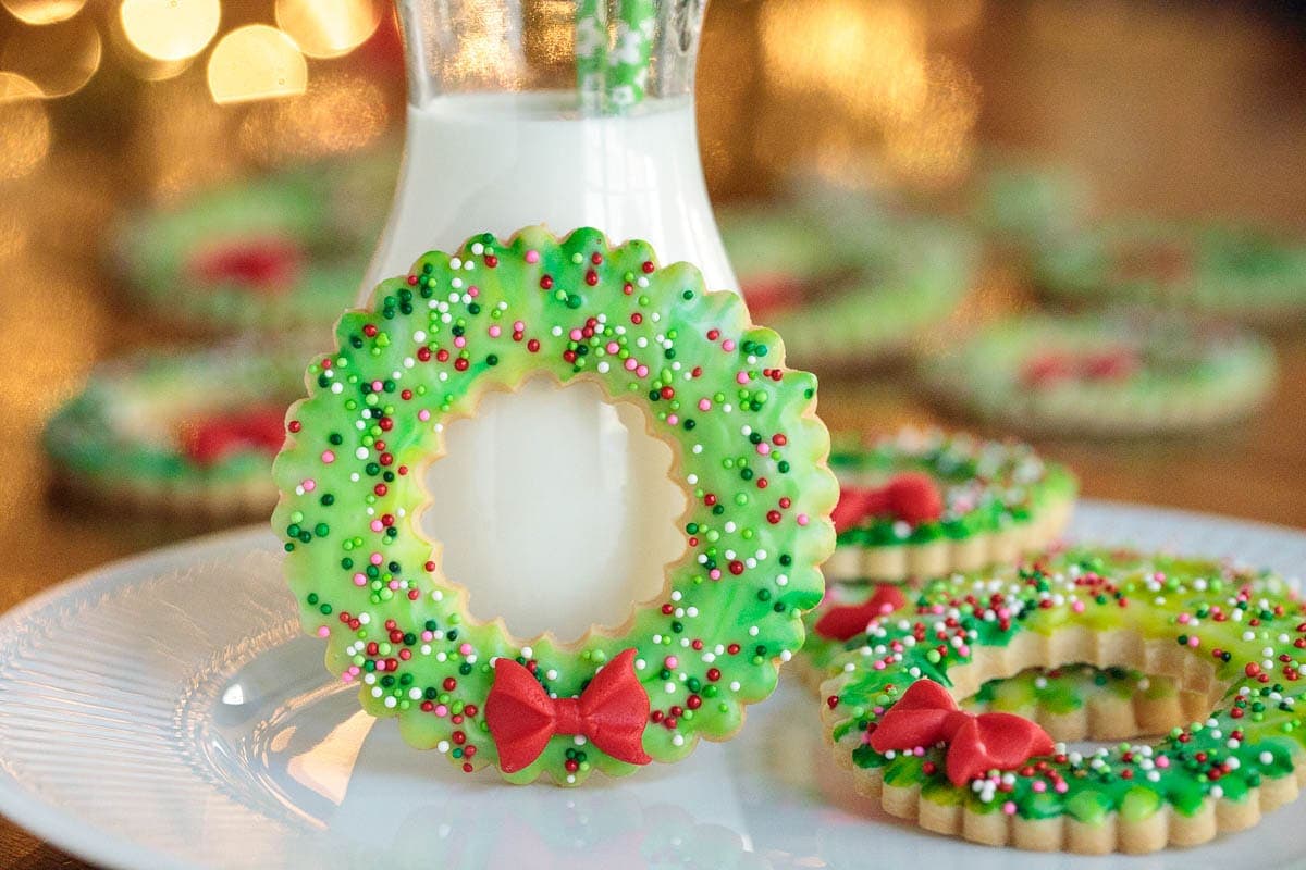 Horizontal photo of a Christmas Wreath Shortbread Cookie leaning up against a jar of milk on a white plate filled with cookies and Christmas lights out of focus in the background.