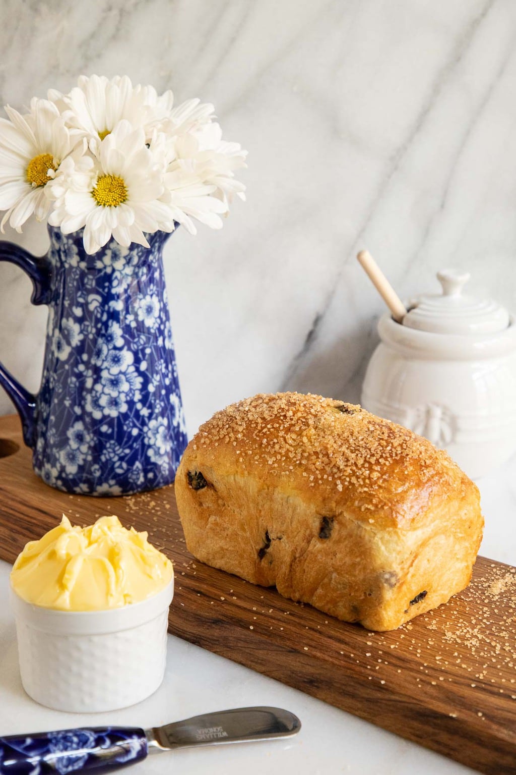 Vertical photo of a loaf of Cinnamon Raisin Brioche Bread on a cutting board with a pitcher of daisies in the background.