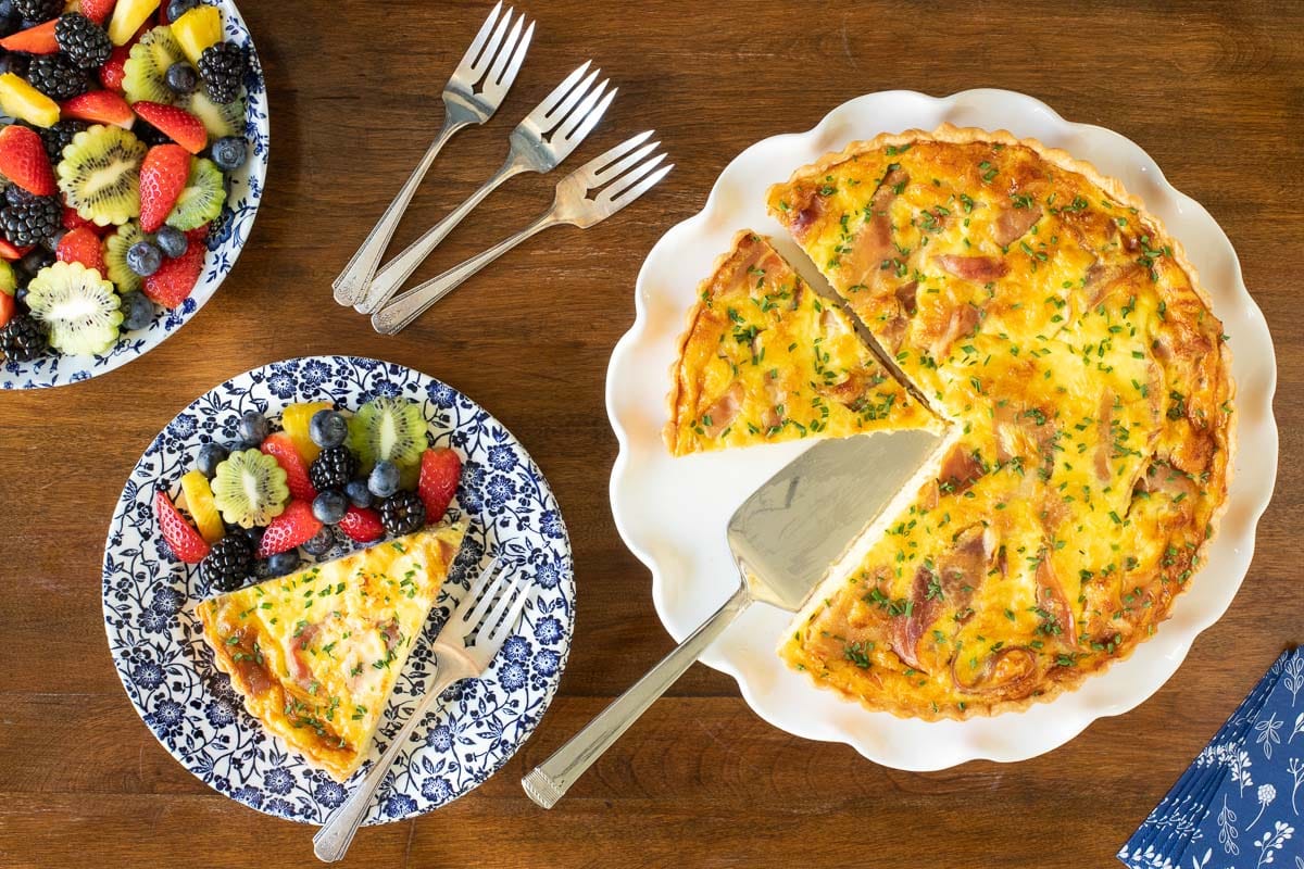 Horizontal overhead photo of a Classic Quiche Lorraine with Prosciutto in a white serving plate and a slice of it on a blue and white patterned platter with a side of fresh fruit.