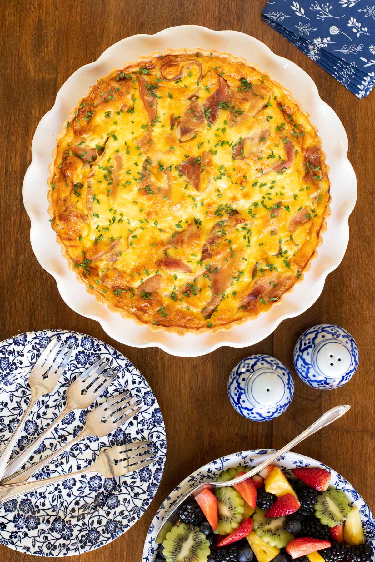Vertical overhead photo of a Classic Quiche Lorraine with Prosciutto on a white scalloped serving plate with a bowl of fresh fruit nearby.